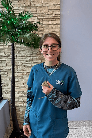 Female wearing glasses and gray scrubs standing in font of a fake lighted palm tree holding a chameleon