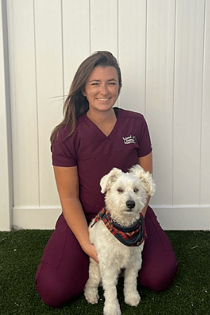 Female with long brown hair wearing maroon scrubs kneeling by white dog