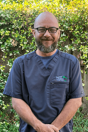 male with beard and shaved head wearing glasses and gray scrubs, arms crossed in front of him