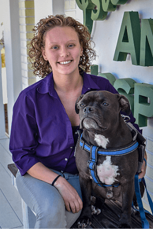 Samantha Scurato female with short blonde curly hair wearing a purple button up shirt posing with a mixed breed black dog